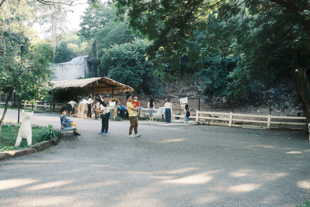Wide park plaza with visitors near a thatched shelter, a white animal statue and dappled shade