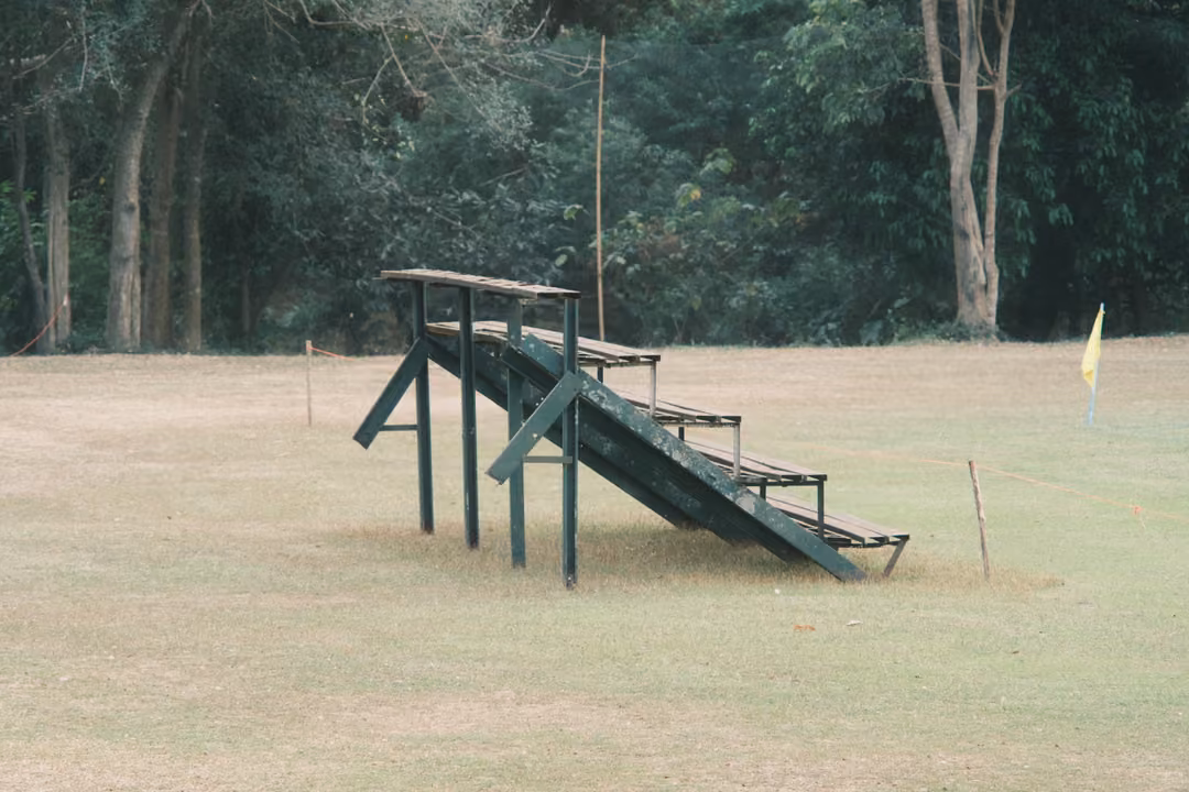 Green wooden bleachers standing alone on a dry grass field, a yellow corner flag and trees behind