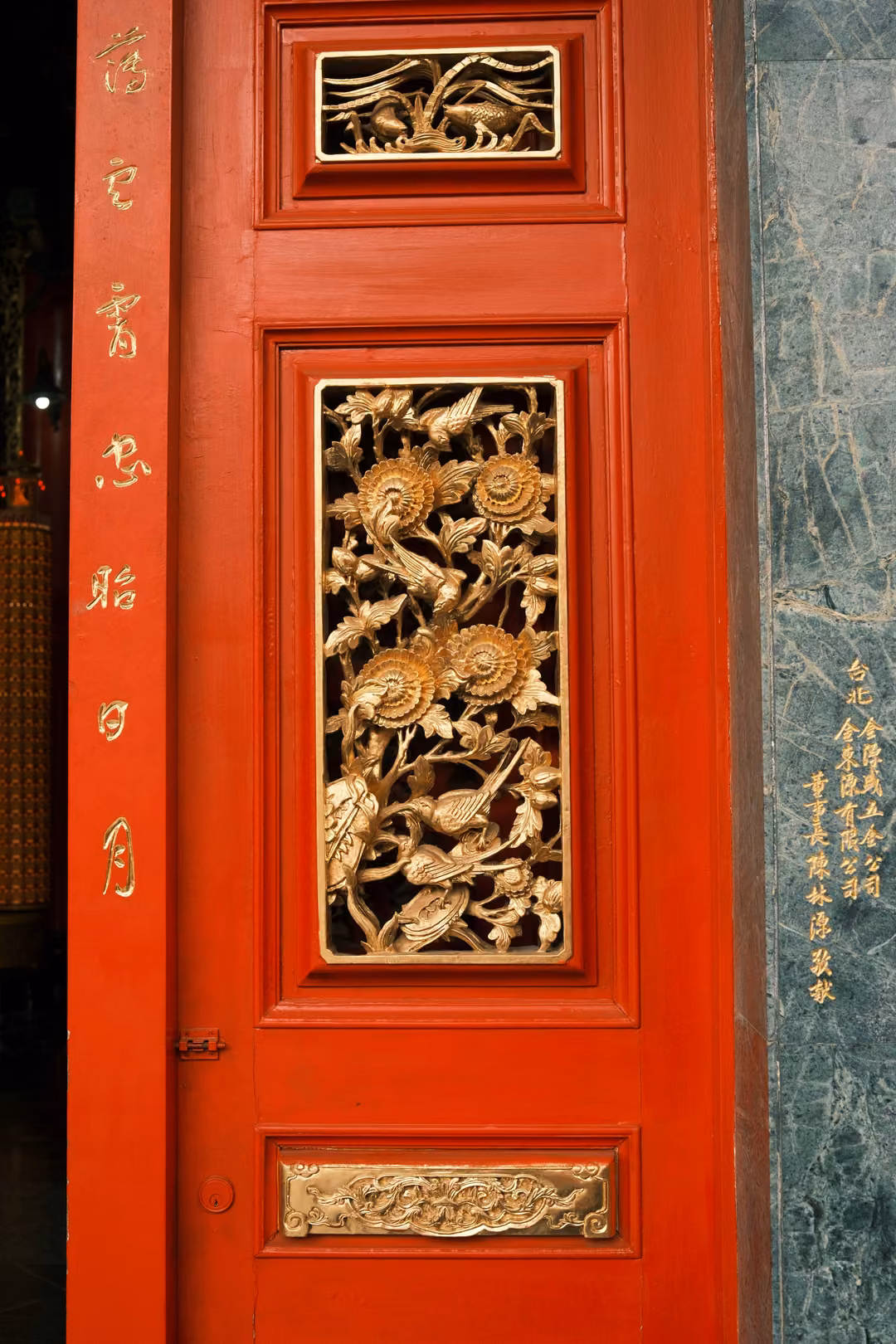 Red temple door with gilded carved panels of flowers and birds, gold characters on its frame, green marble beside