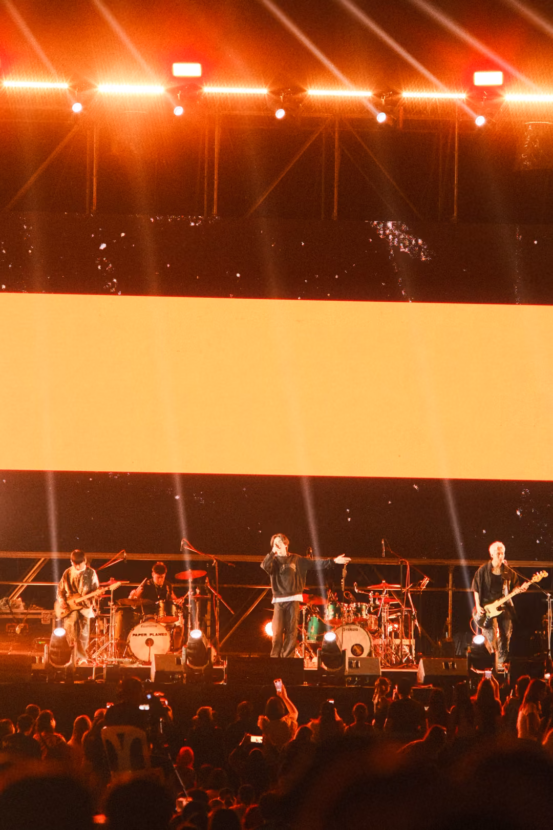 Vertical view of a stage glowing orange with a bright pale band of light across the screen, band and crowd below.