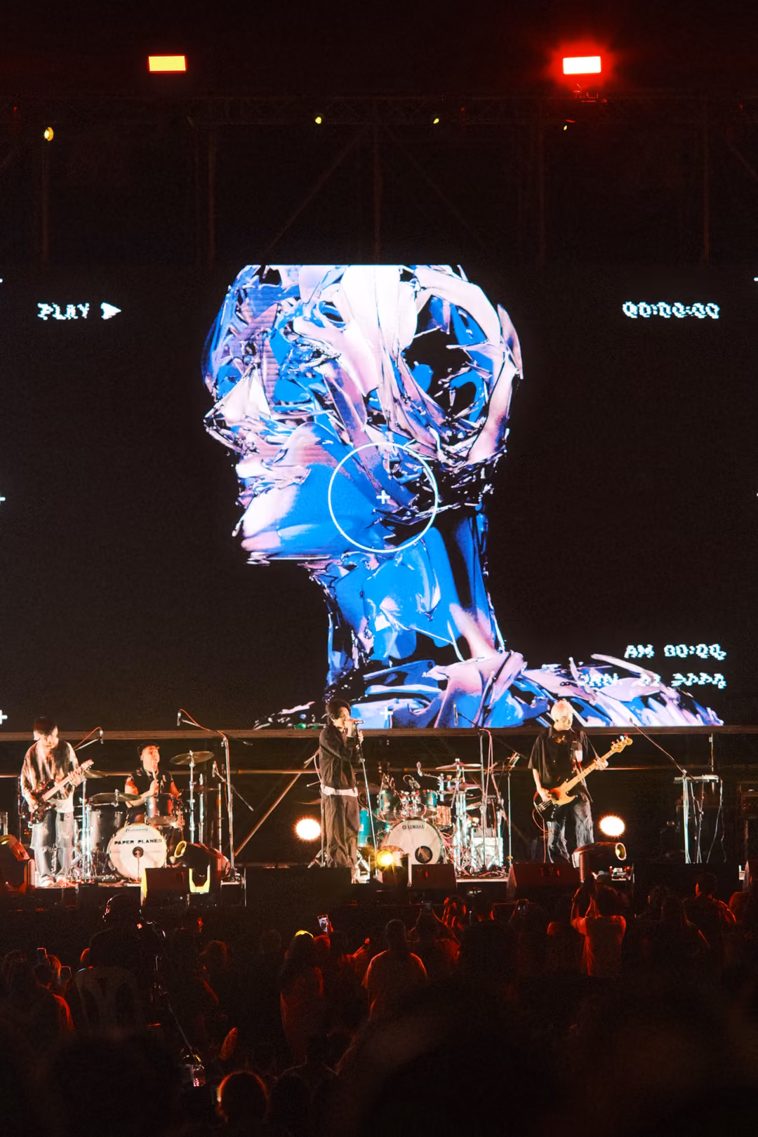 Vertical view of a screen showing a blue and white sculpted head with a PLAY label, band performing below.