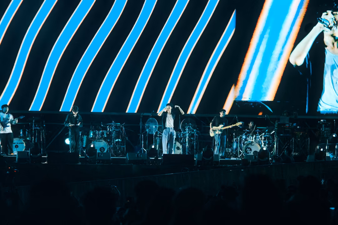 Band on stage below a screen of slanted blue and white stripes, the singer's arm visible on screen at right.