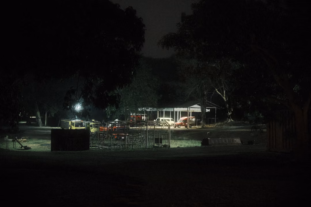 Distant field at night with parked cars and a covered shelter lit by a single lamp, dark trees around