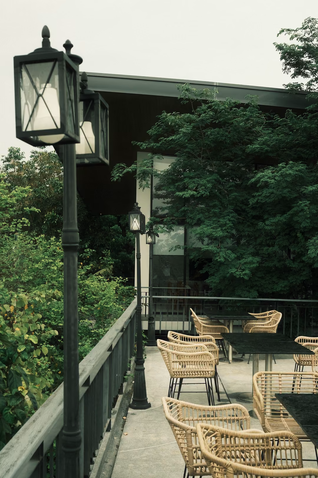 Upright view of a terrace lamp post with lantern tops, wicker chairs along the railing and trees beyond.