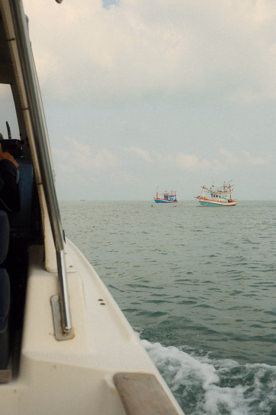 The boat's white hull cuts through water with foam, two fishing boats ahead on the sea.