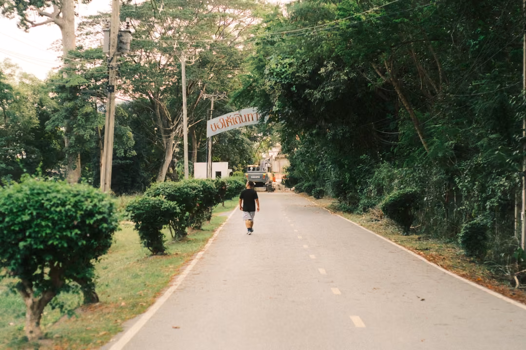 A person in a black T-shirt walks along a shaded road toward an excavator and a roadside arch sign.