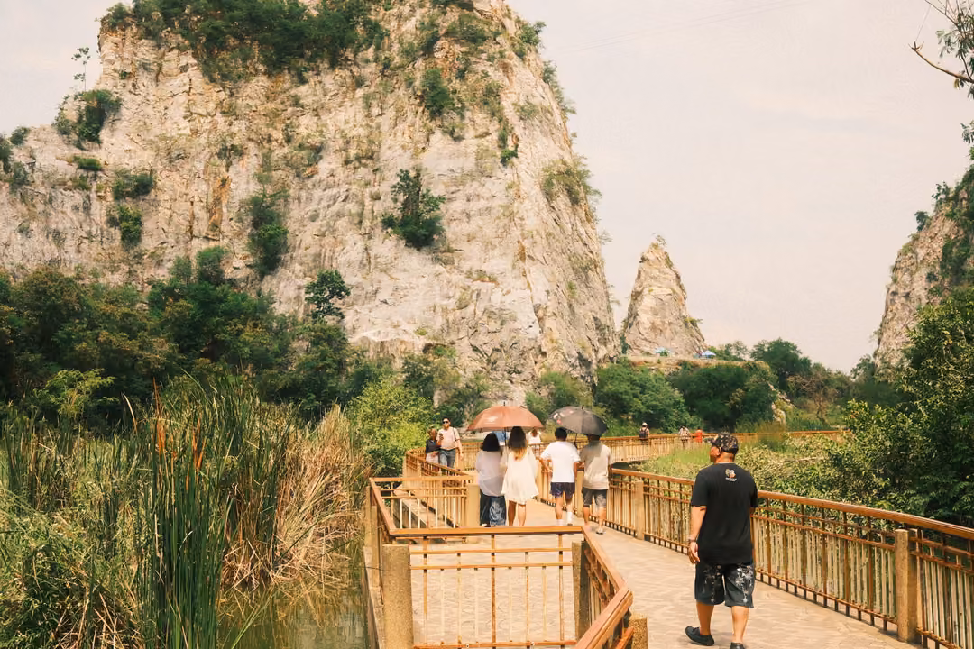 Lakeside boardwalk with visitors under umbrellas, a man in black walking, limestone cliffs and a rock spire behind