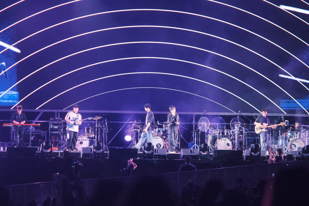 Musicians on stage under glowing white arcs on a violet screen, two vocalists standing together at centre.