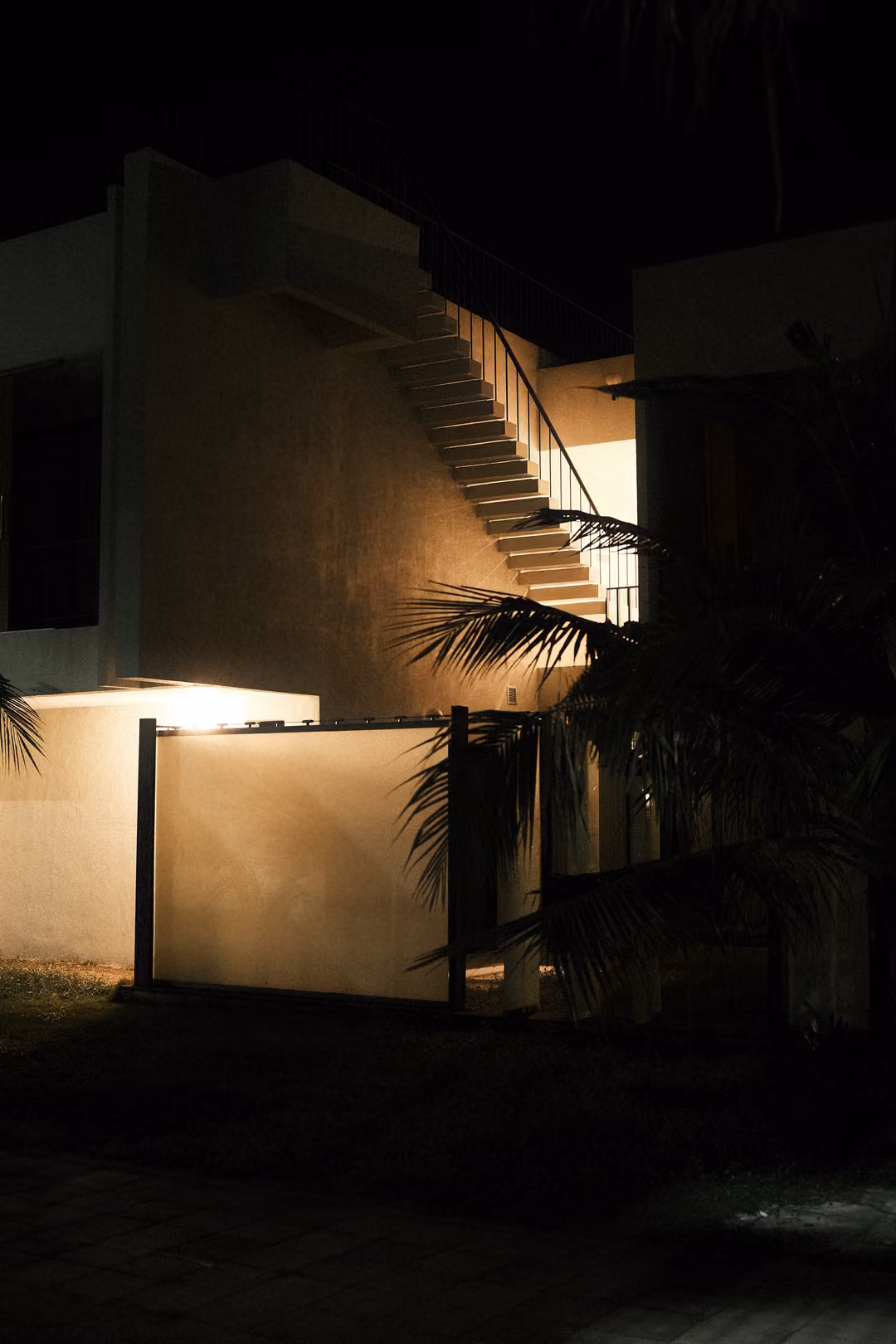 Side of a white villa at night with an outdoor staircase and wall lit warm, palms in shadow