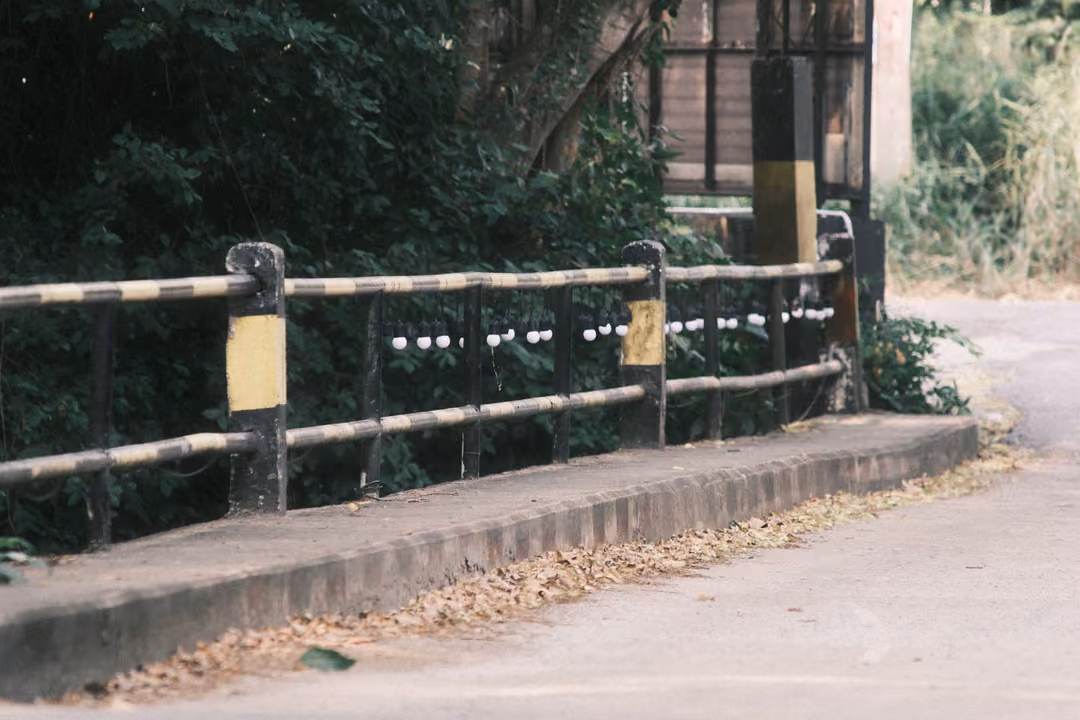 A low concrete bridge railing with yellow and black posts beside a road edge strewn with dry leaves.