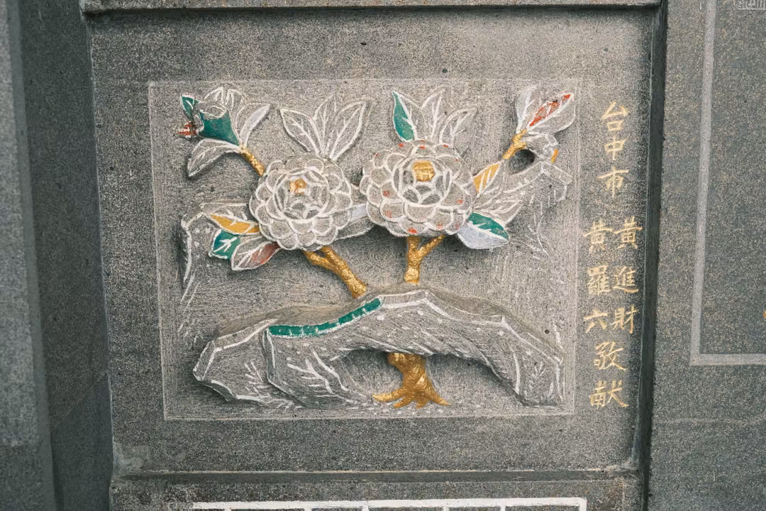 Stone relief of two large blossoms on a branch over a rock, with green leaves and gold inscription