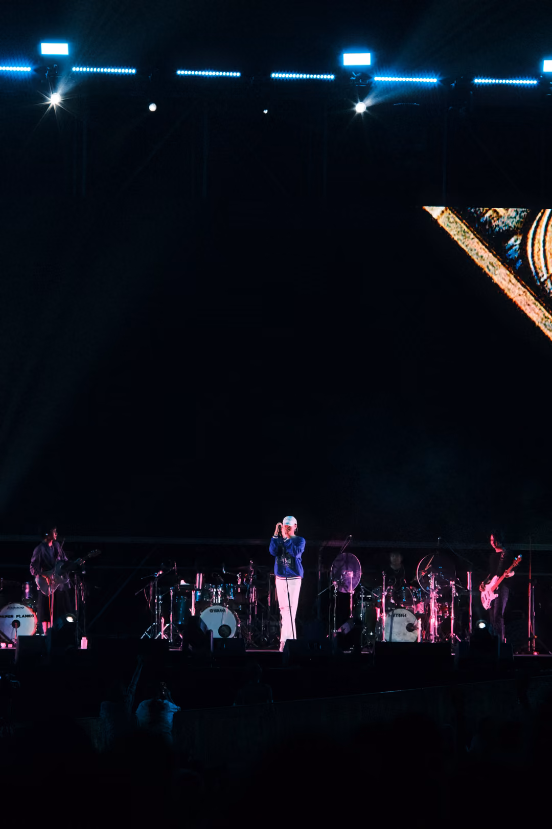 Vertical view of a dark stage, the singer in a blue jacket at centre, guitarists either side, a gold frame corner above.