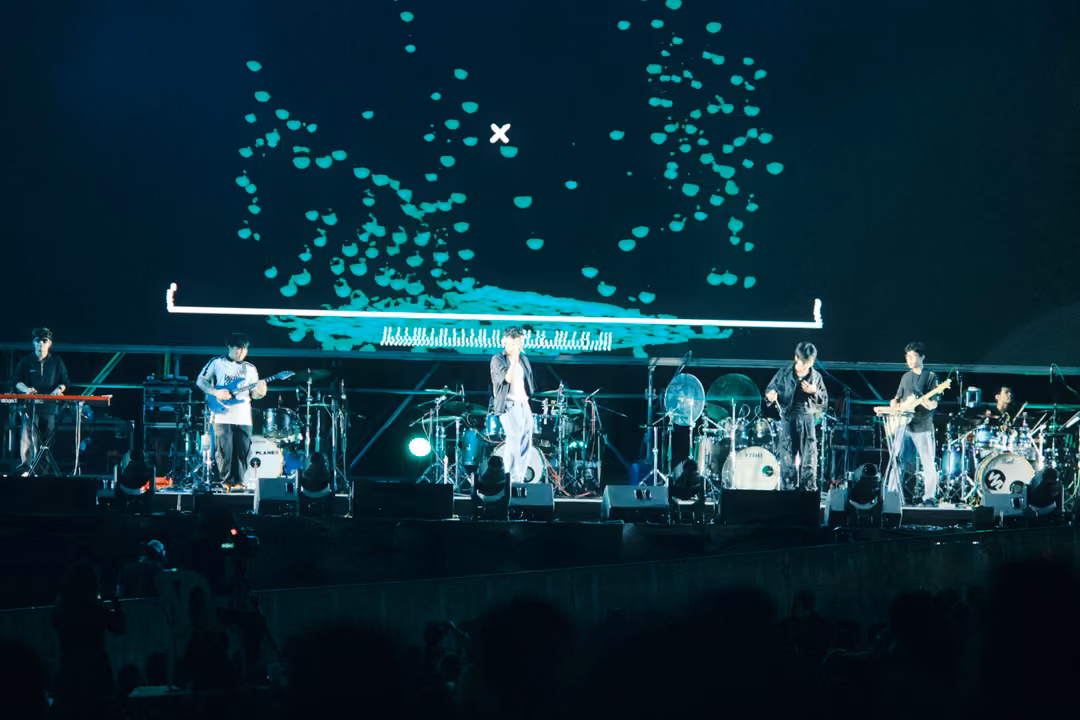 Musicians on a dark stage under a screen filled with drifting turquoise shapes above a white bracket.