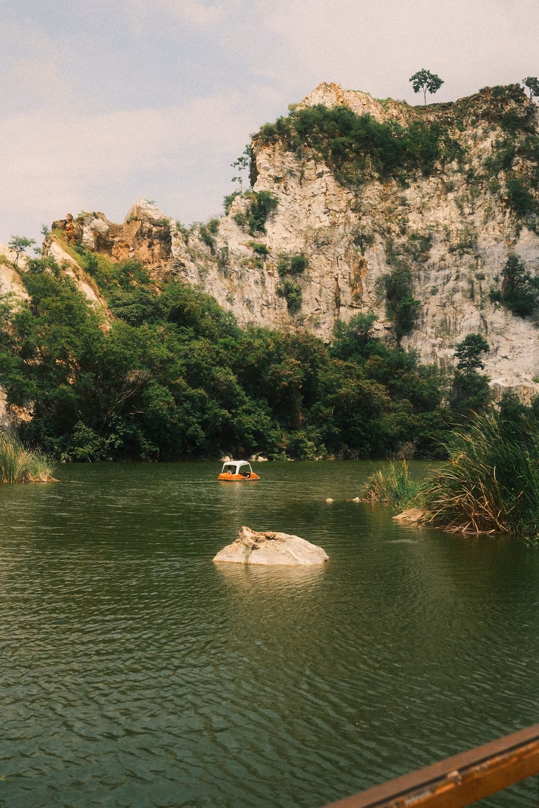 Green lake with a small pale rock island and a pedal boat beneath a limestone cliff and partly cloudy sky