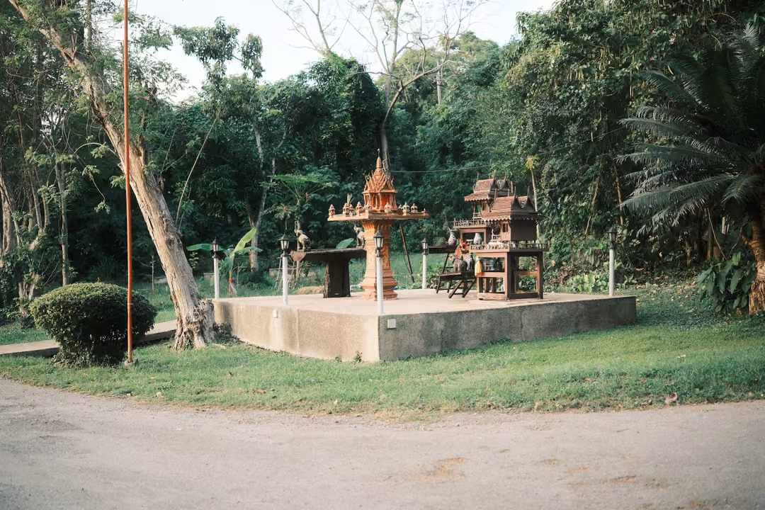Ornate orange spirit house and wooden shrine on a concrete platform among trees, a tall orange pole beside