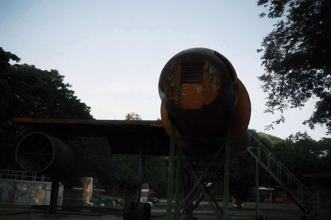 Silhouette of a rusted aircraft fuselage end on a metal stand with stairs, trees behind, pale sky