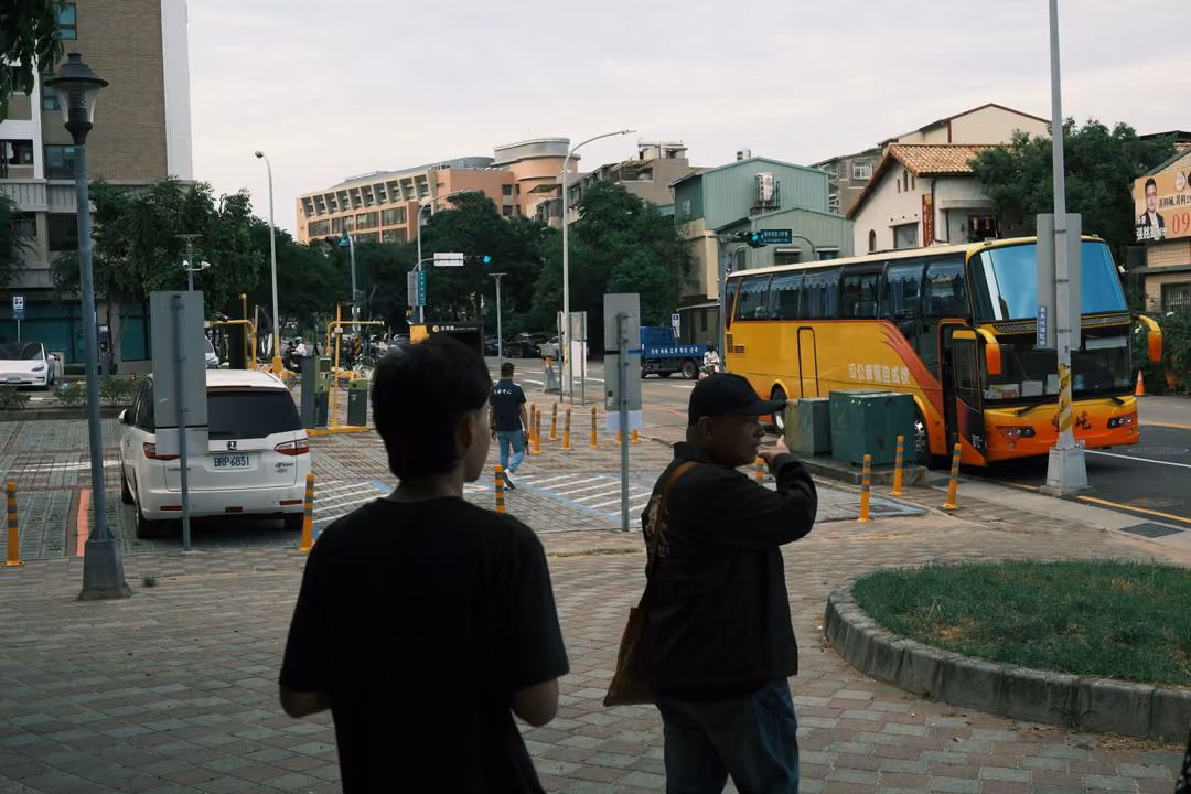 Two people seen from behind on a paved plaza, one filming a street where a yellow tour bus is parked