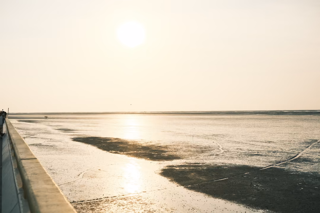A low sun over wide, shining mudflats beside the yellow railing of a long causeway