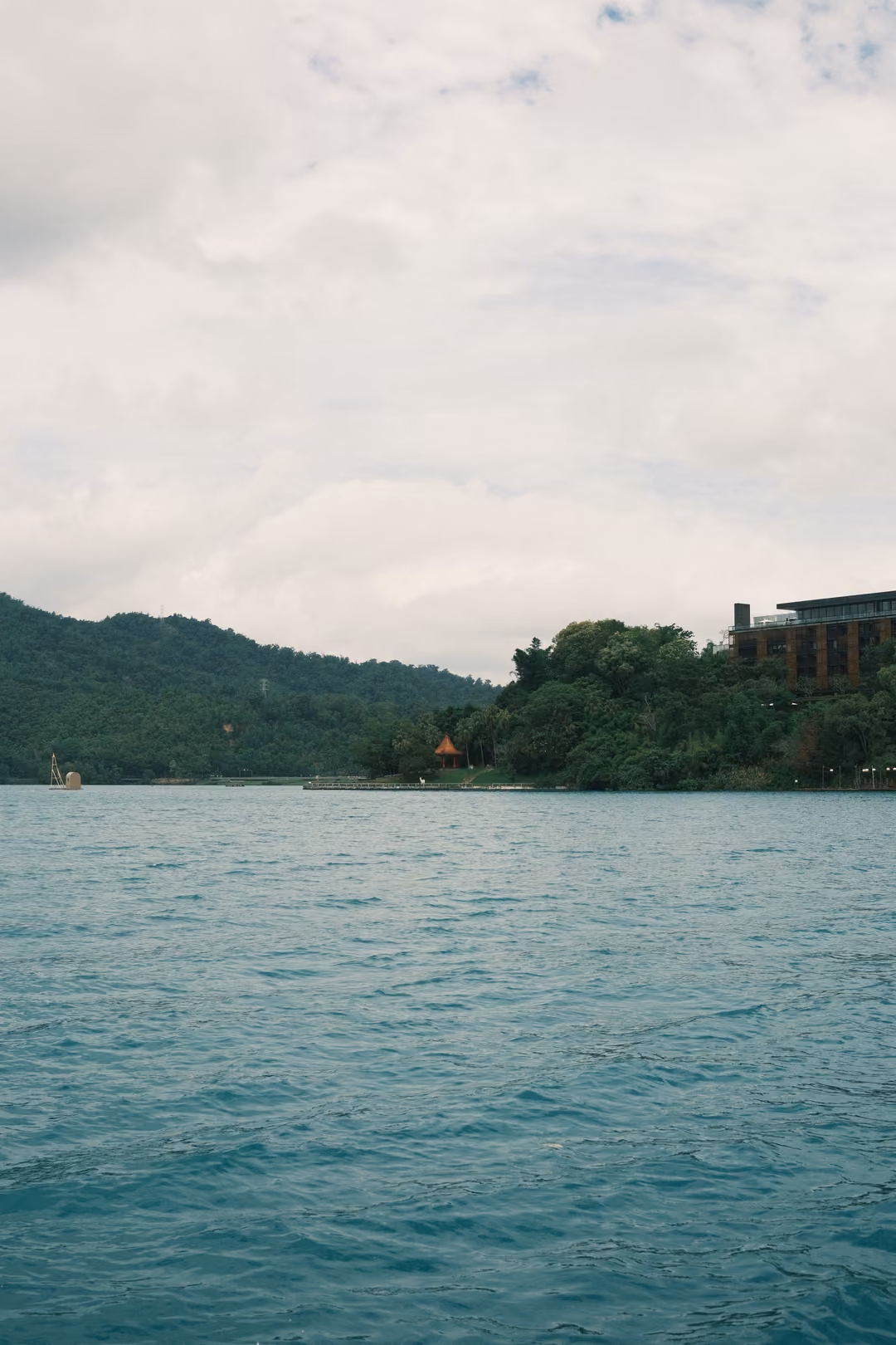 Expanse of teal lake water under a pale overcast sky, a wooded headland and buildings at the right