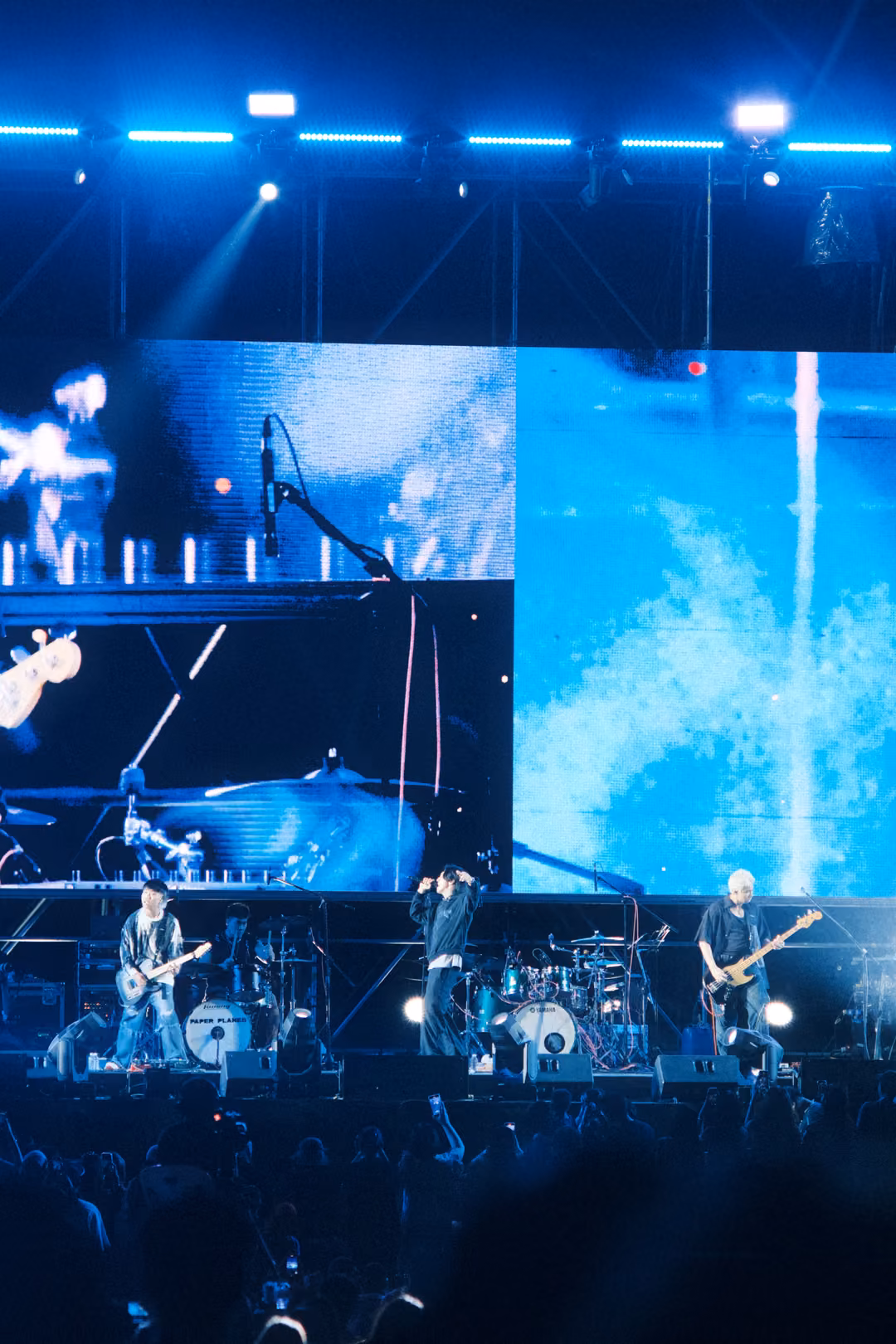 Vertical view of a stage with blue-lit screen panels, a singer at centre with one arm raised, two guitarists beside.