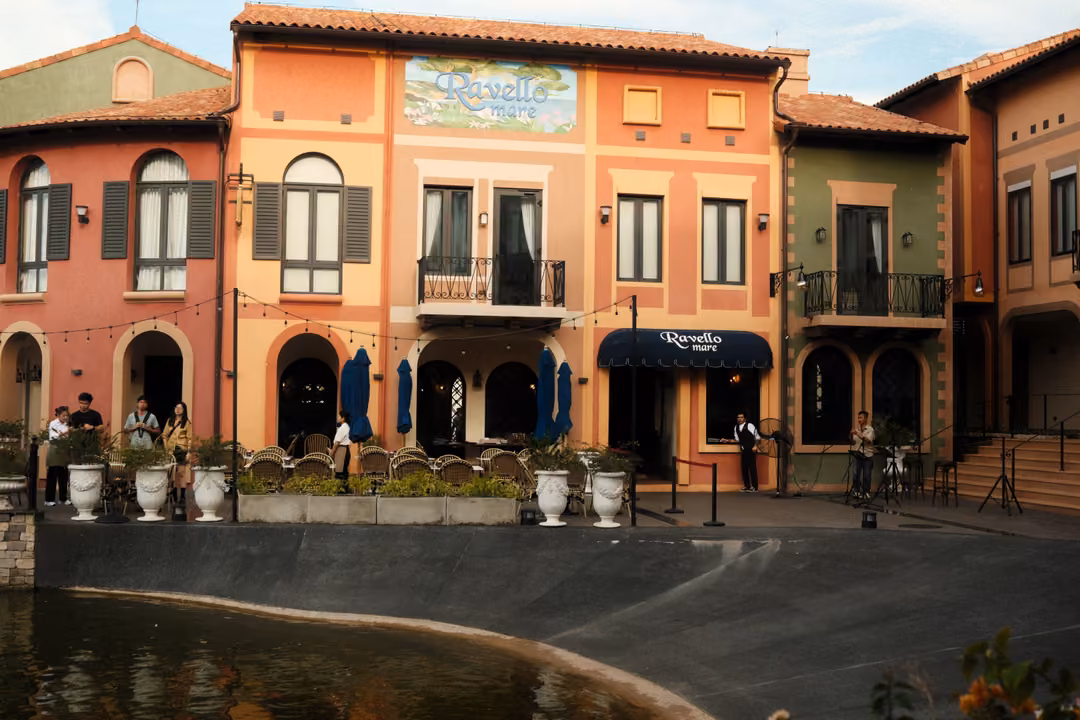 Pink and orange facades with a painted Ravello sign, navy umbrellas and people by a curved pond edge