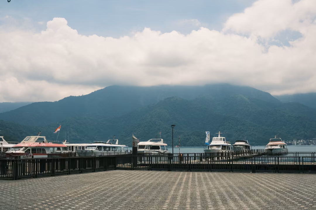 Empty tiled plaza leading to a pier with white boats on a lake, mountains under bright cumulus clouds