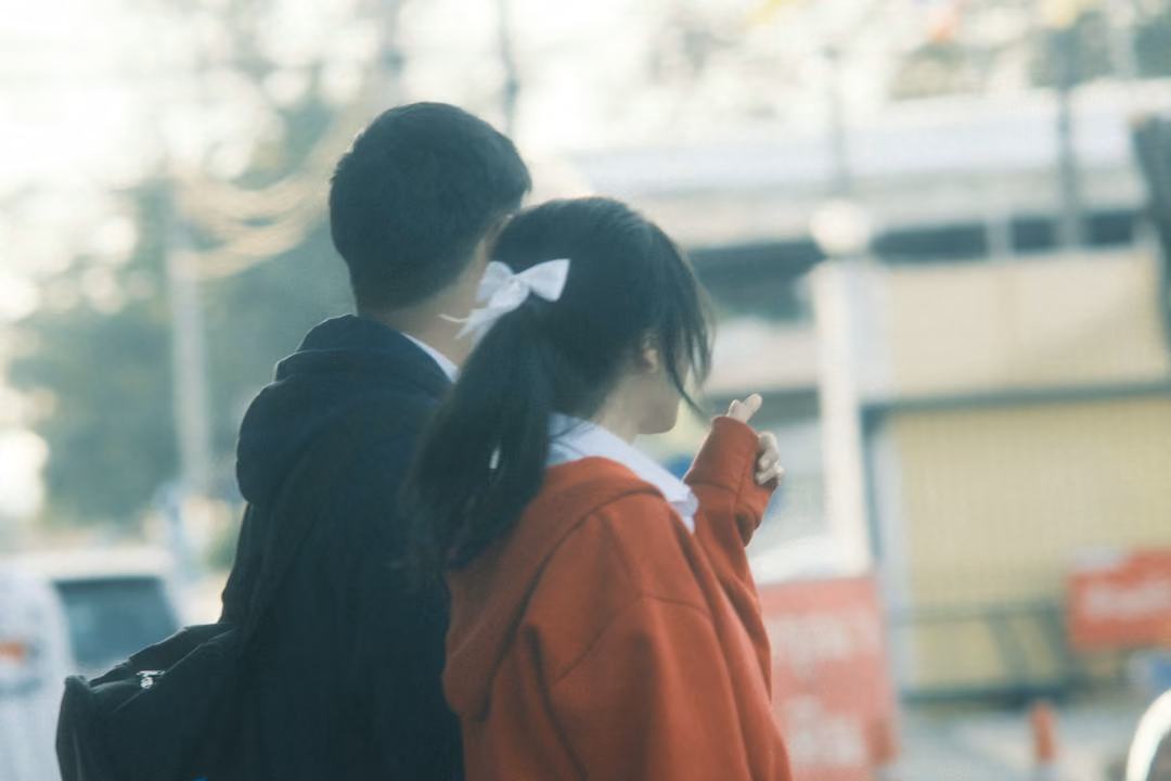 Two students seen from behind, one in a red jacket with a white hair bow pointing ahead, the other in a dark hoodie.