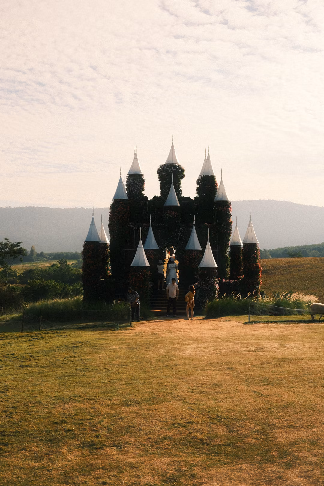 A castle-shaped structure covered in flowers and greenery, with white conical spires, visitors at its steps.