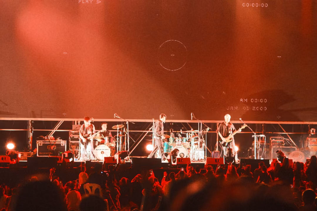 Band on a red-lit stage below a hazy red screen with a faint crosshair and AM 00:00 date stamp, crowd in red light.