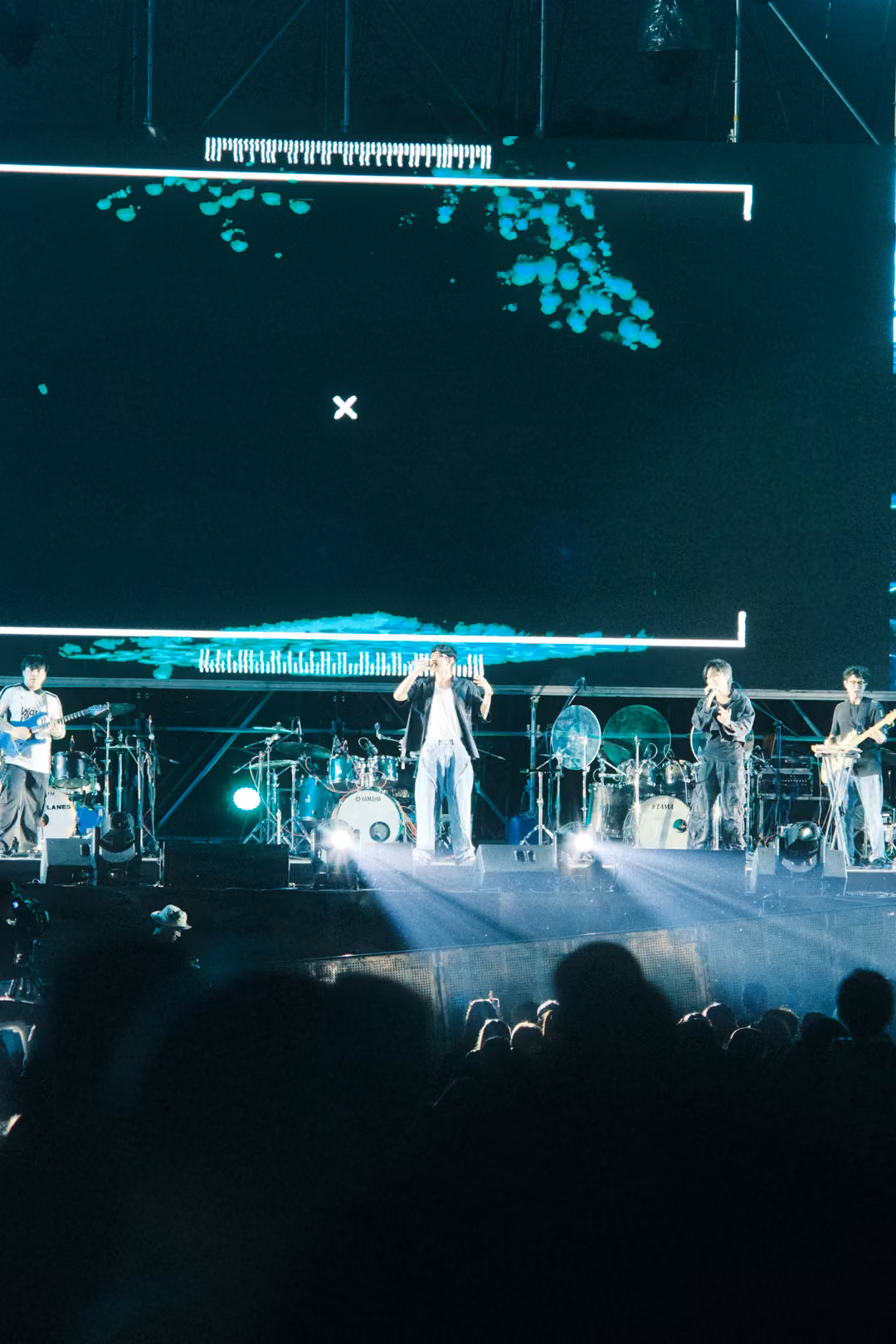 Singer at centre stage under a white spotlight beam, a few turquoise shapes drifting at the top of the screen.