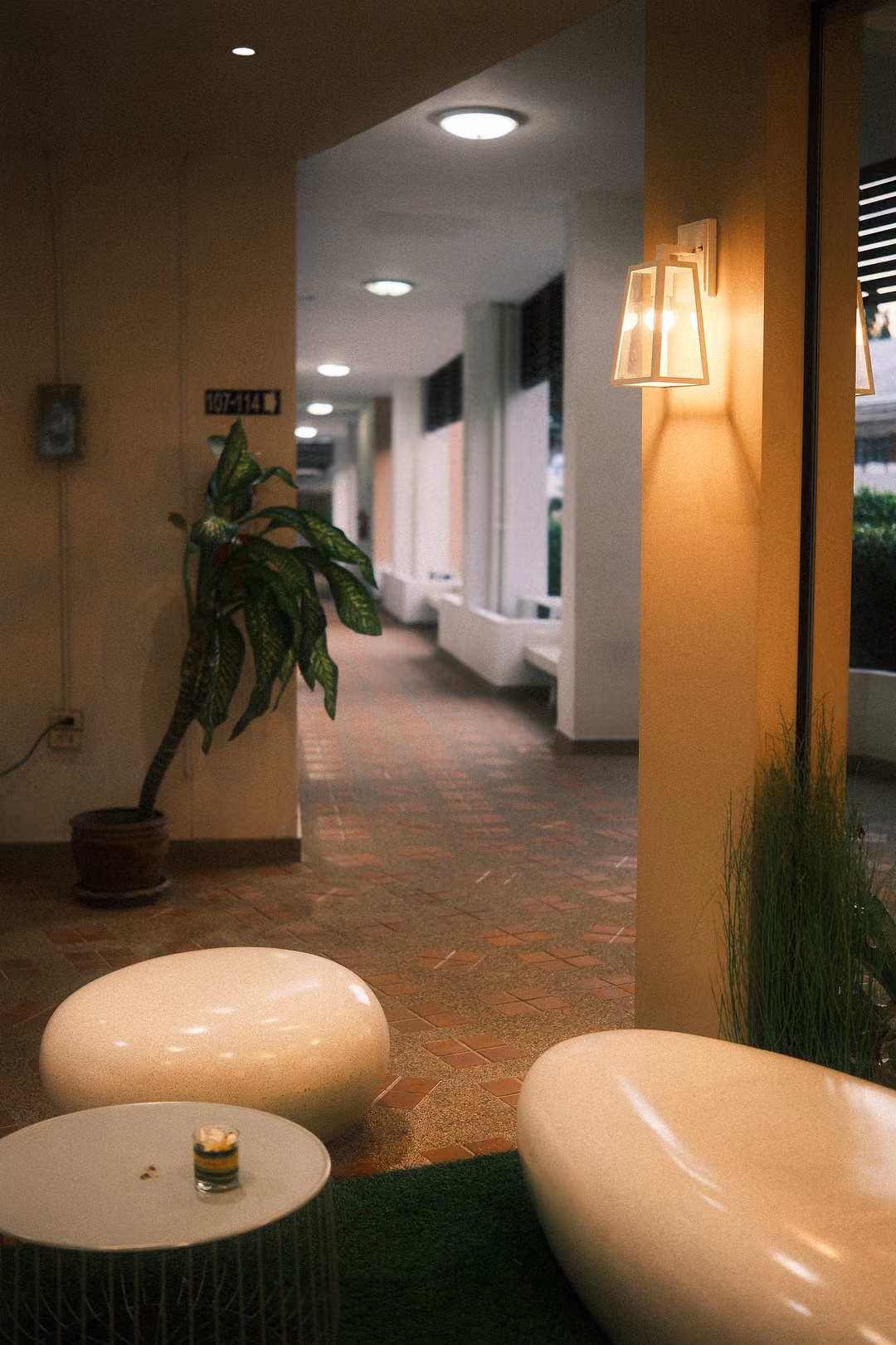 Warmly lit hotel corridor with a wall lamp, potted plant, white pebble seats and a small candle on a table