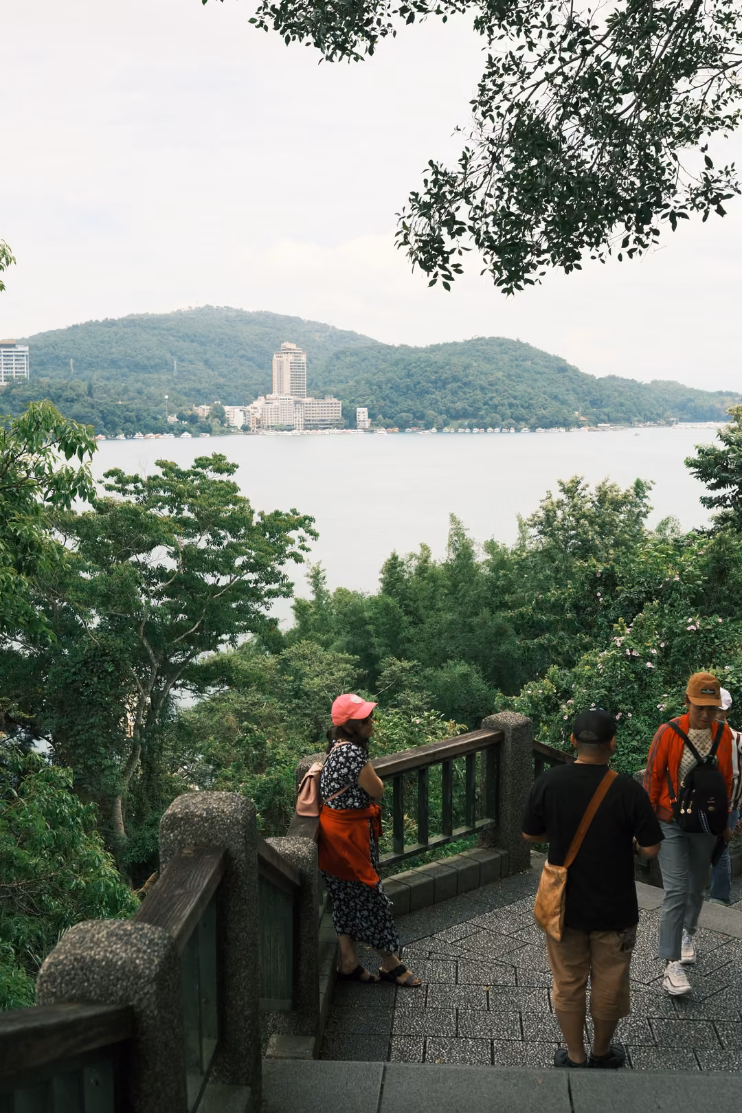Visitors at a stone railing on a hillside landing look out toward a lake and a tall building on the far shore
