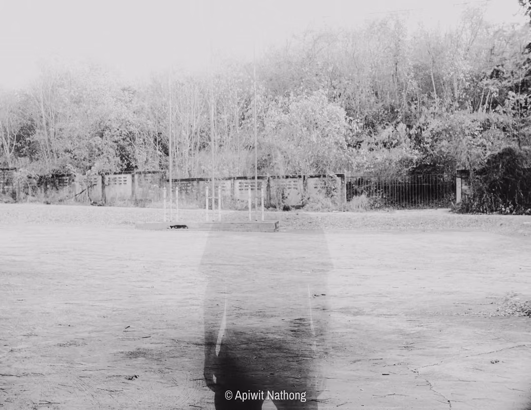 Black and white ghostly translucent figure fading into a bare yard, fence and pale trees behind