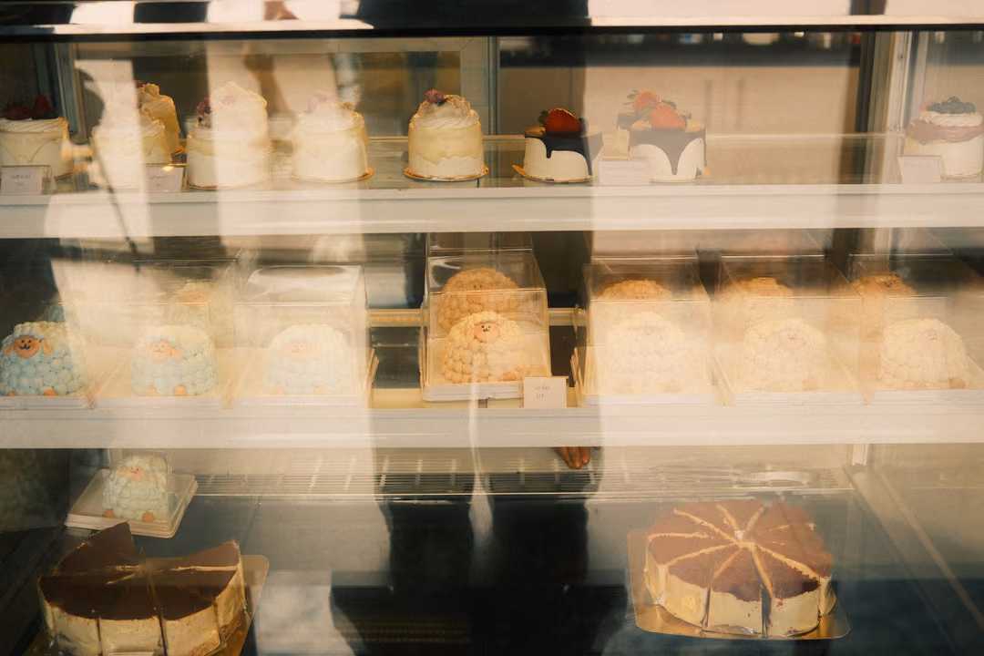 A glass display case filled with rows of cakes, its surface catching reflections of the room.