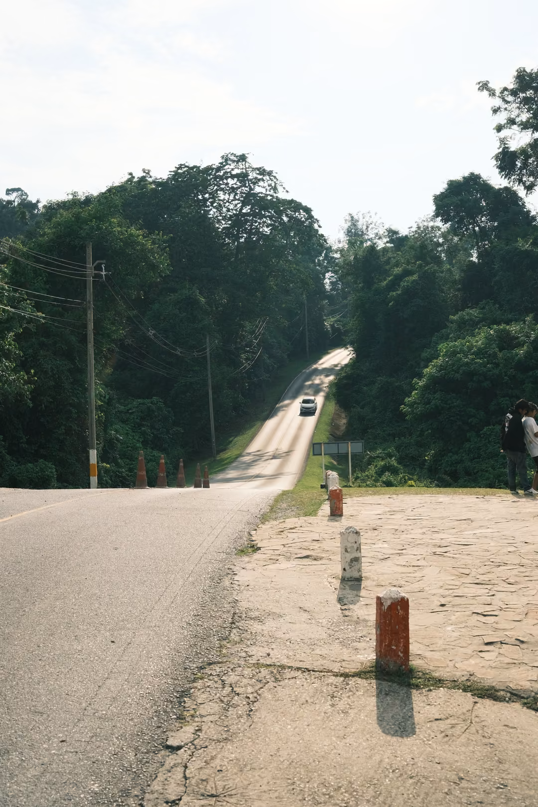 The same steep road with a car descending toward the posts and a person standing at the right edge.