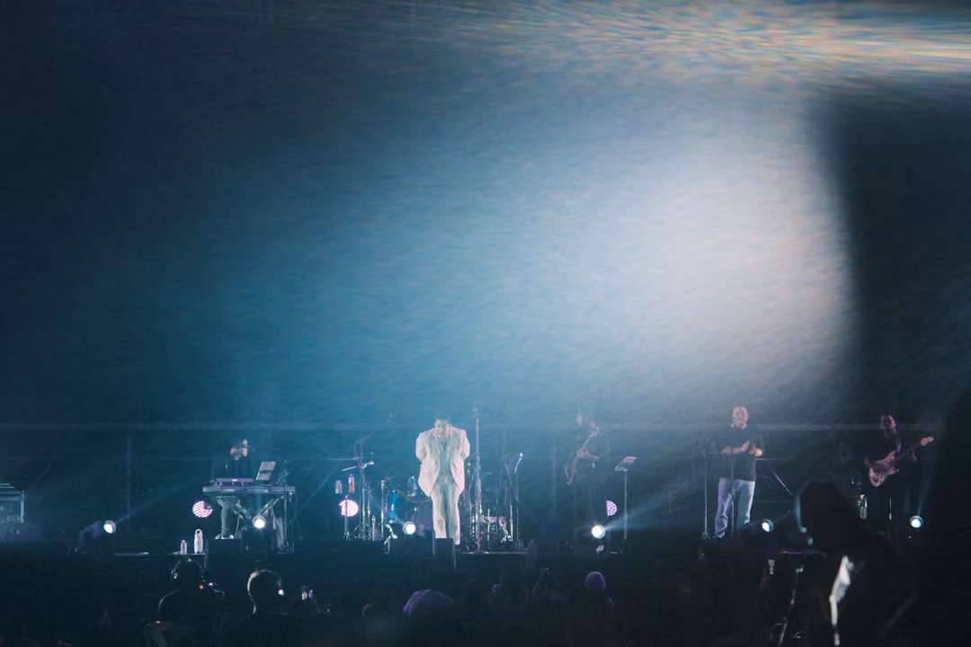Stage washed in white haze and soft light, a singer in a white suit centre stage, band and keyboards around him