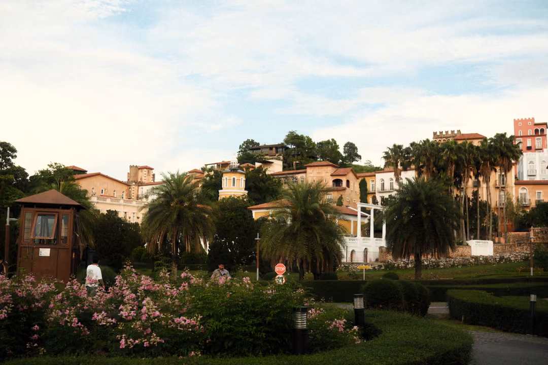 Hillside of pastel buildings above palm trees, clipped hedges, a small wooden guard hut and pink flowers