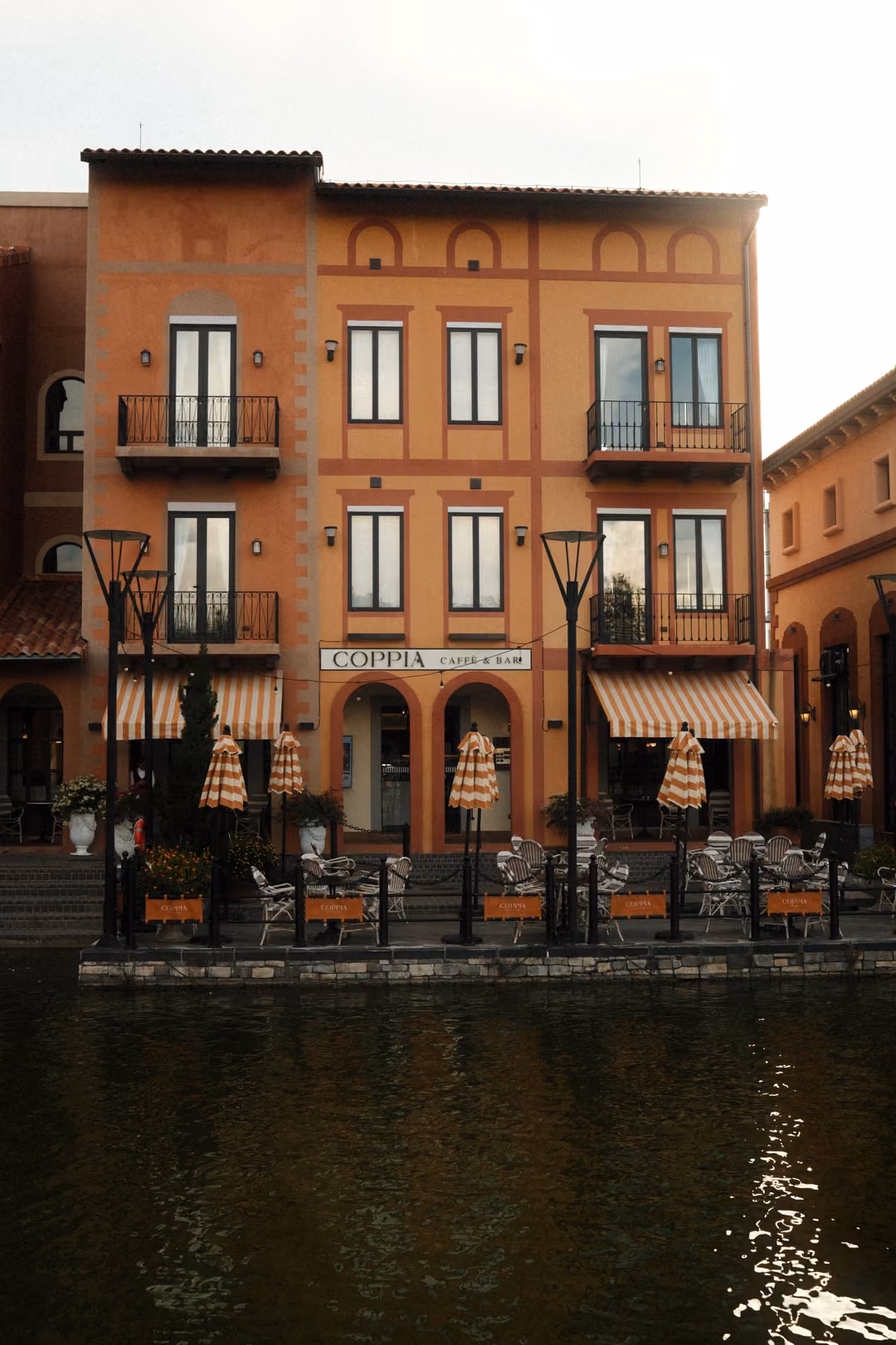 Orange building with balconies and striped closed umbrellas on a waterside terrace, dark water in front