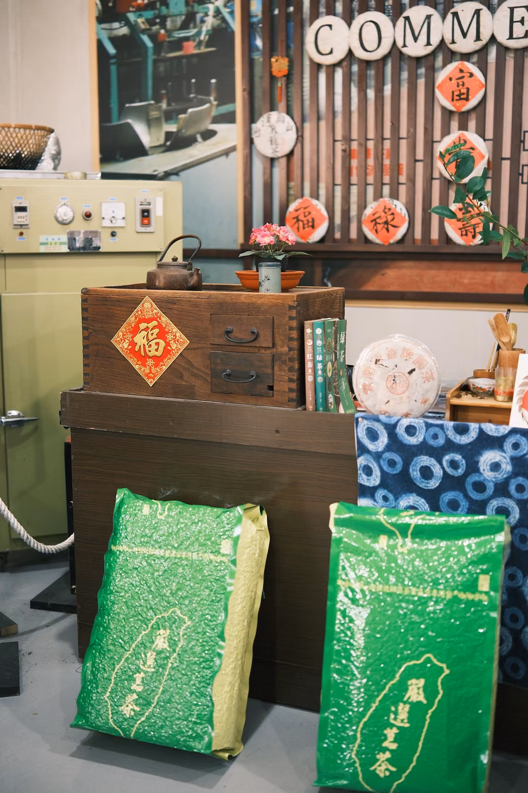 Green bags of tea leaning against a wooden counter with a red 福 diamond, a teapot and potted flowers on top