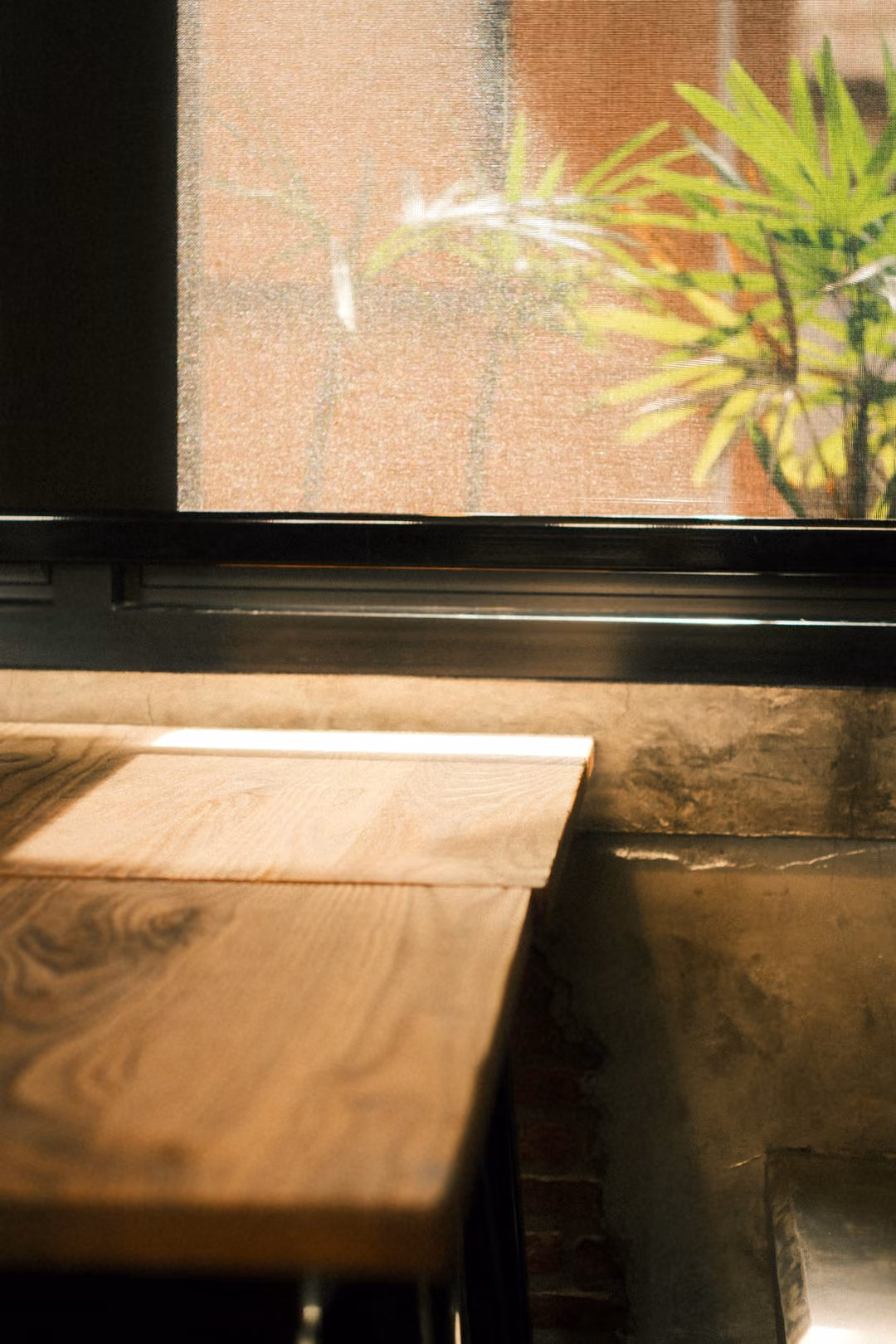 An empty wooden table against a concrete wall below a black-framed window with green plants outside