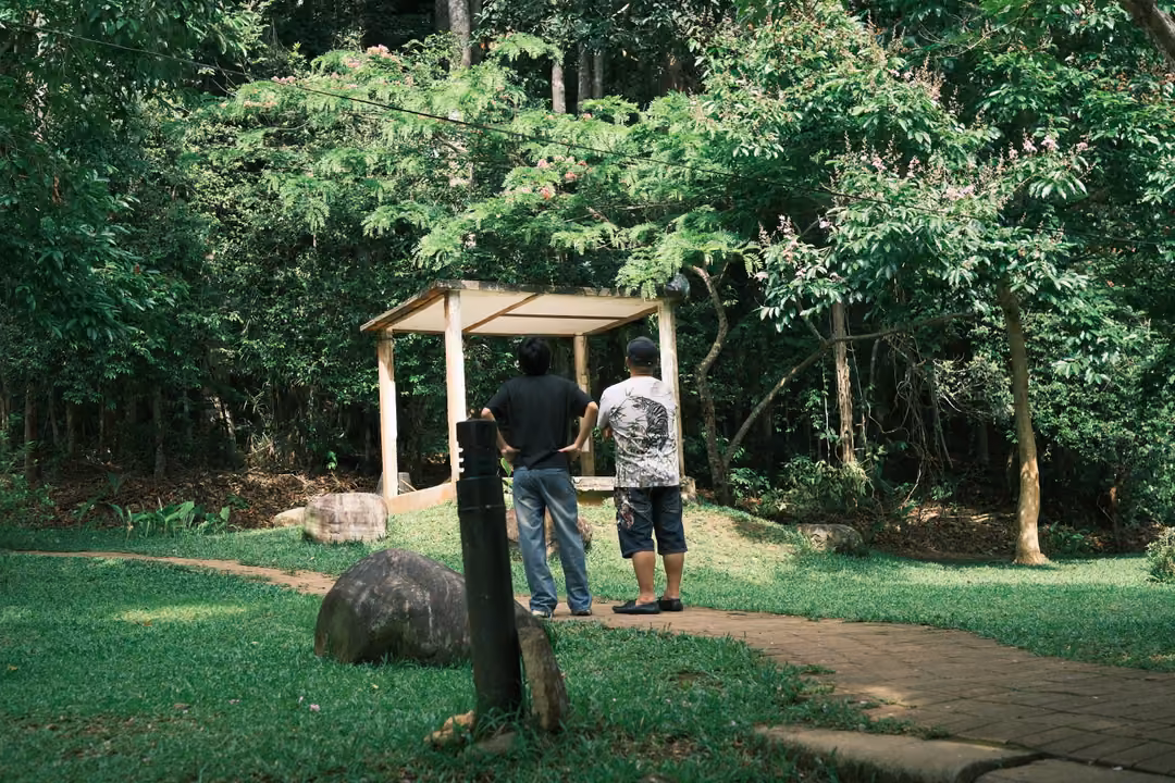 Two people standing by a small wooden shelter at the edge of a forest, a dark post in front
