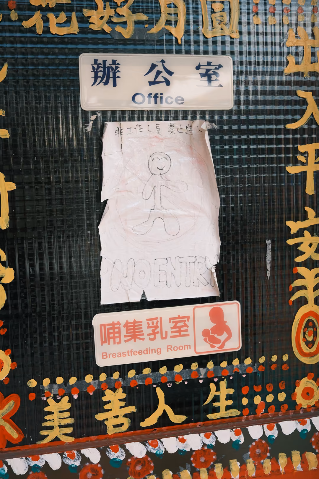 Glass door with an Office sign, a Breastfeeding Room sign, a taped child's drawing and gold painted characters