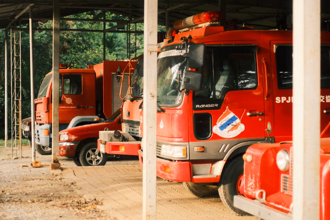 Red fire engines parked under an open-sided shelter, with steel posts in front and an emblem on a truck door.