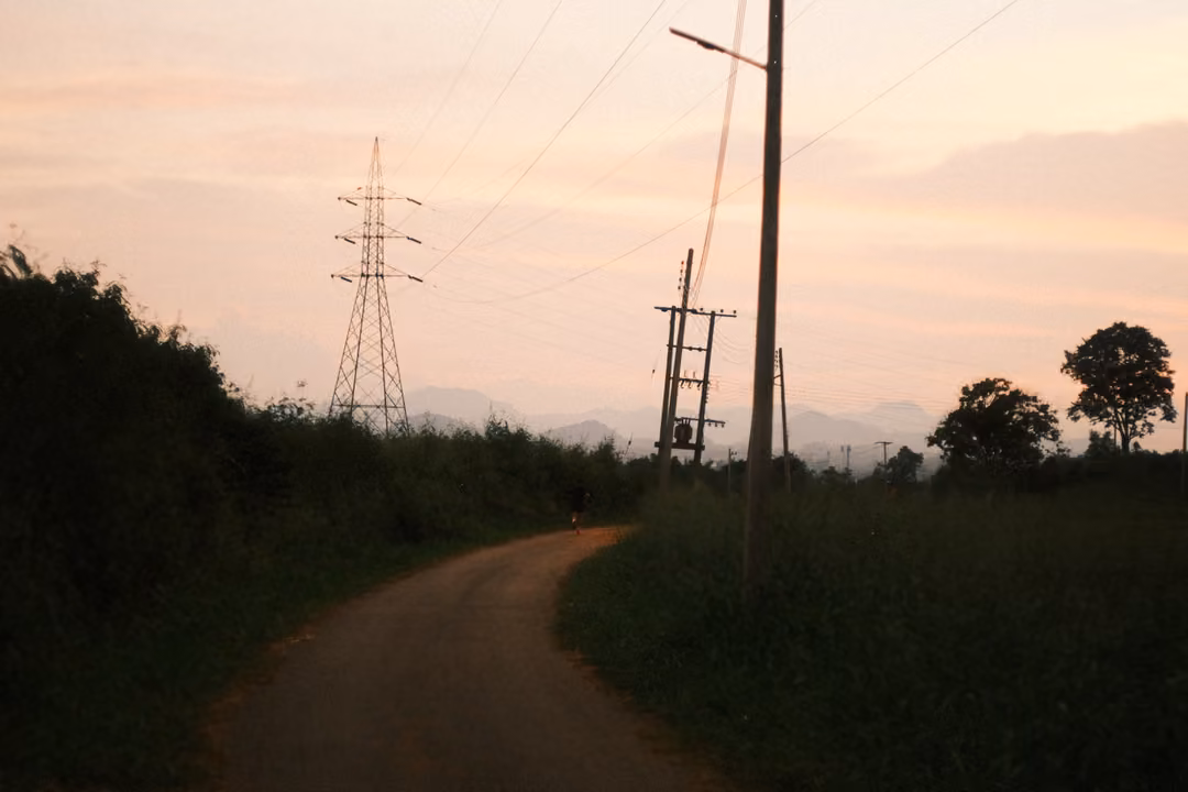 Narrow road curving past a pylon and power poles under a pink evening sky, mountains in the distance