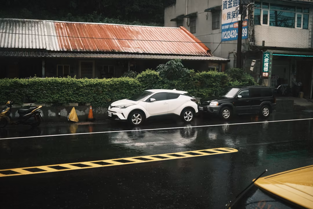 Rainy roadside with a red-roofed shed, hedge, and white and black cars, a yellow car hood at lower right