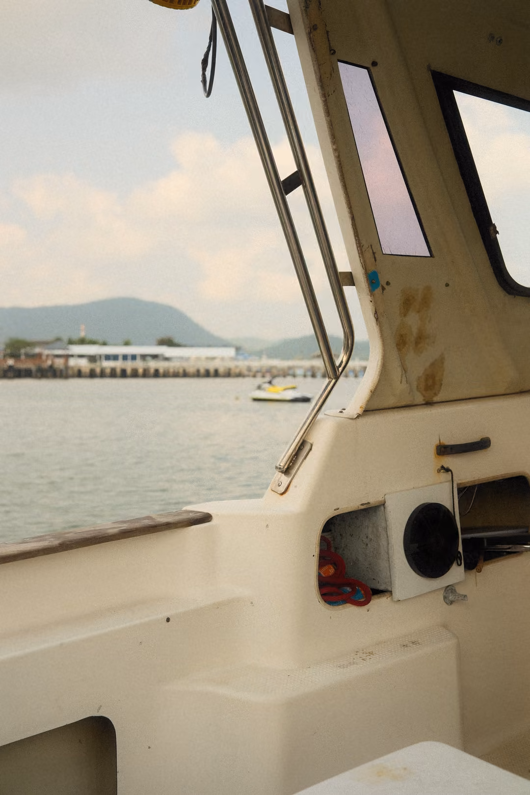 View past a boat's cabin frame to a long pier and a green hill, a jet ski on the water nearby.