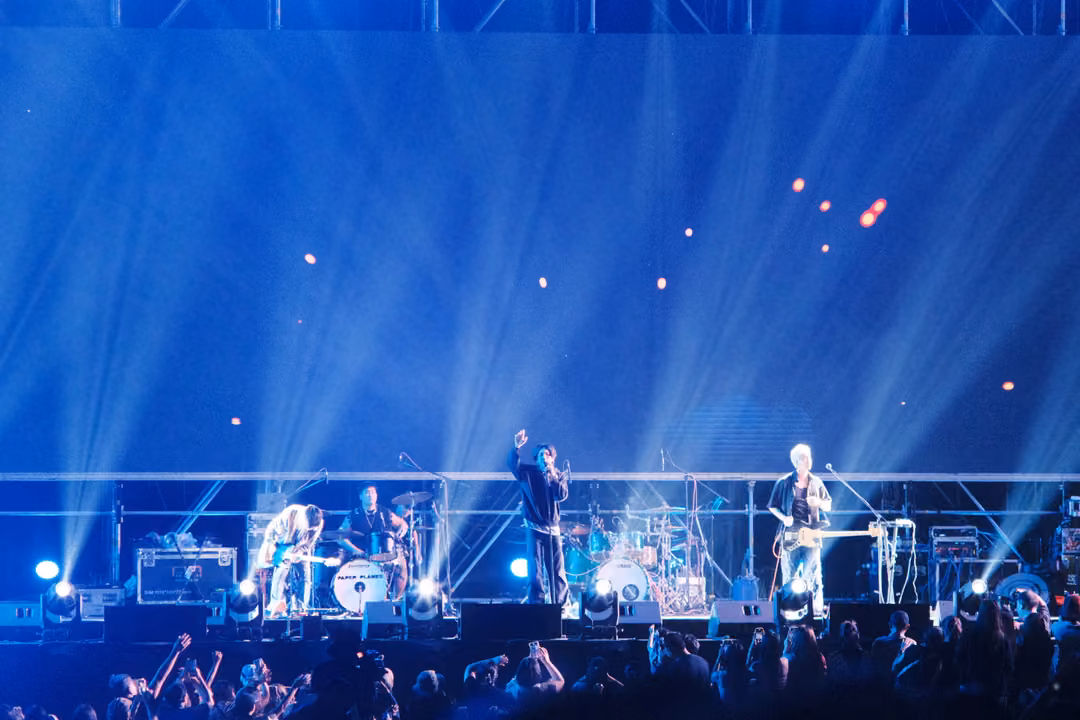 Band performing beneath blue light beams and drifting orange sparks, crowd with raised arms in the foreground.