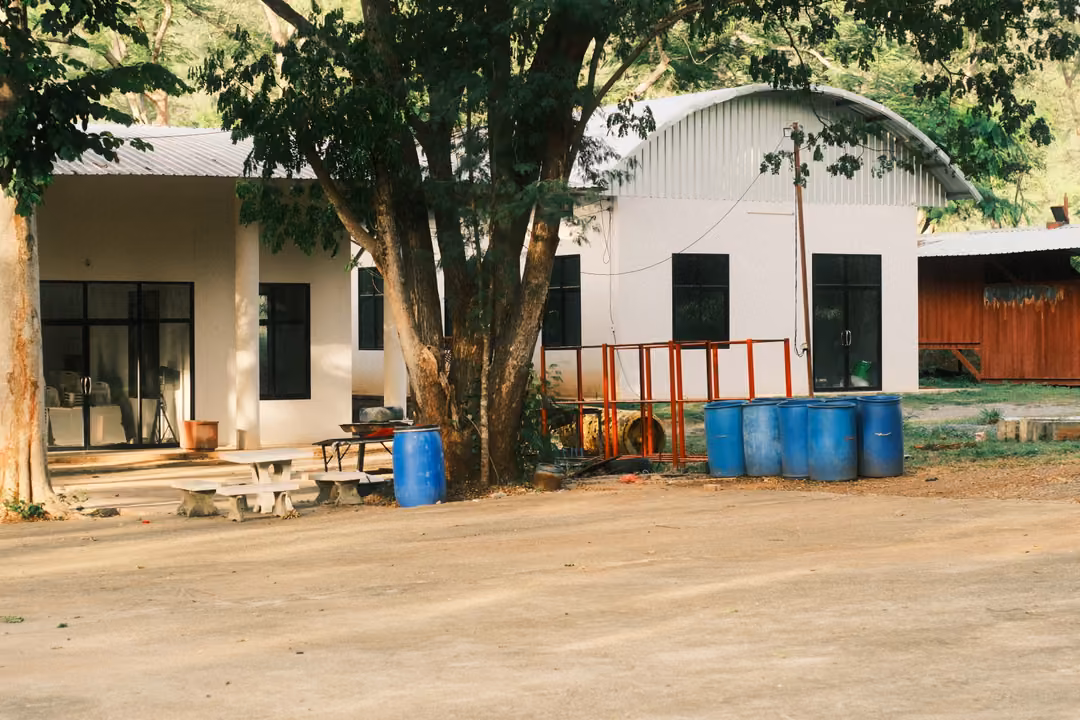 A white building with a curved metal roof, blue plastic barrels, an orange railing and picnic benches beneath a tree.
