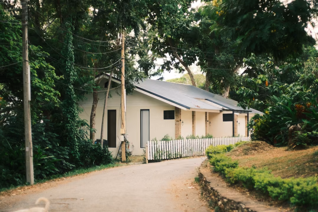 A paved road curving past a white cottage with a grey roof and white picket fence, trees and power poles around.