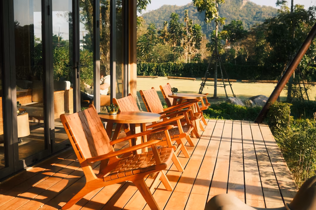 Wooden deck in low sunlight with slatted lounge chairs and small tables, garden and wooded hill beyond